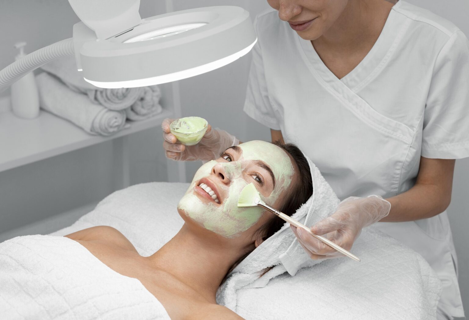 Esthetician applying brightening facial mask in a well-lit spa room.